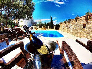 Zona junto a la piscina con sillones de mimbre, un muro de piedra y una botella de champán en un balde.