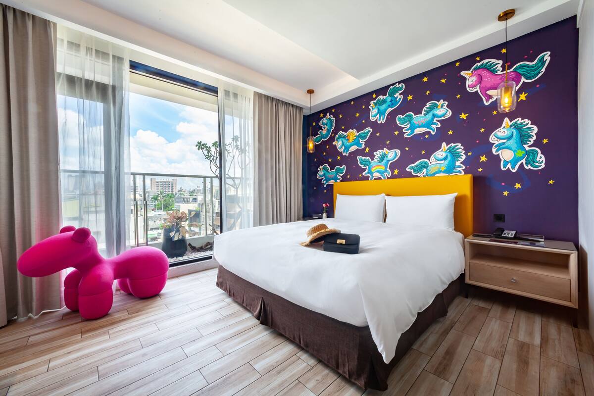 Una habitación de hotel moderna con una cama grande, una mesita de noche, un mural llamativo en la pared y vistas a la ciudad.