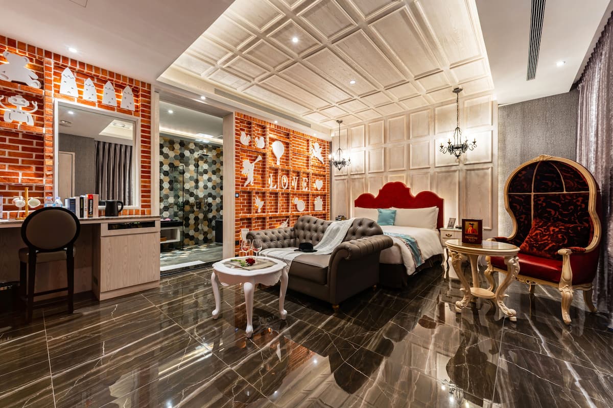 Una habitación de hotel moderna con una cama grande, un espacio de estar con un sofá y sillones, una pequeña mesa de comedor y una zona de cocina.