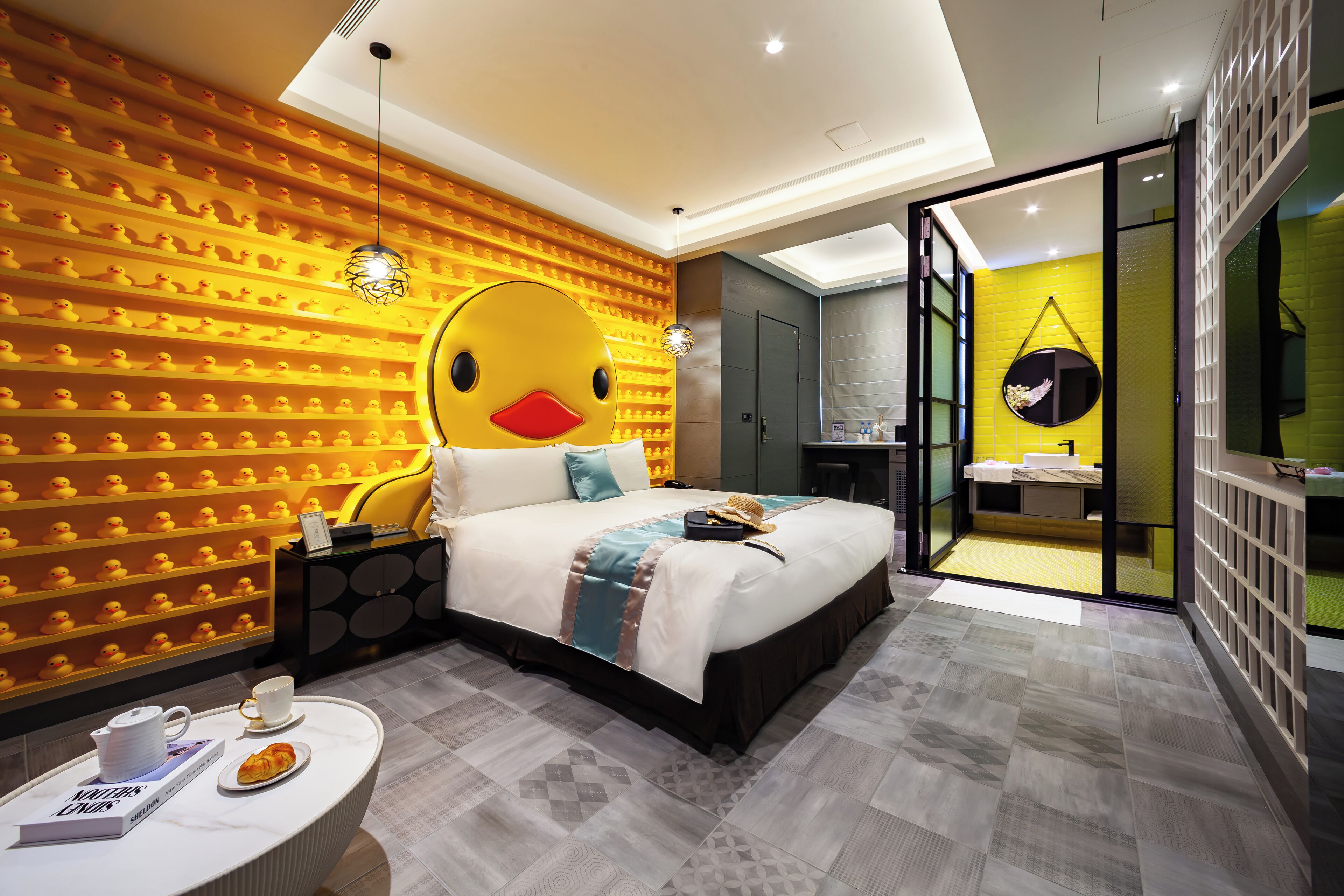 Ein modernes Hotelzimmer mit einer auffälligen Wand im Enten-Design, einem großen Bett, einem Nachttisch mit Buch und Tasse sowie Blick ins Badezimmer.
