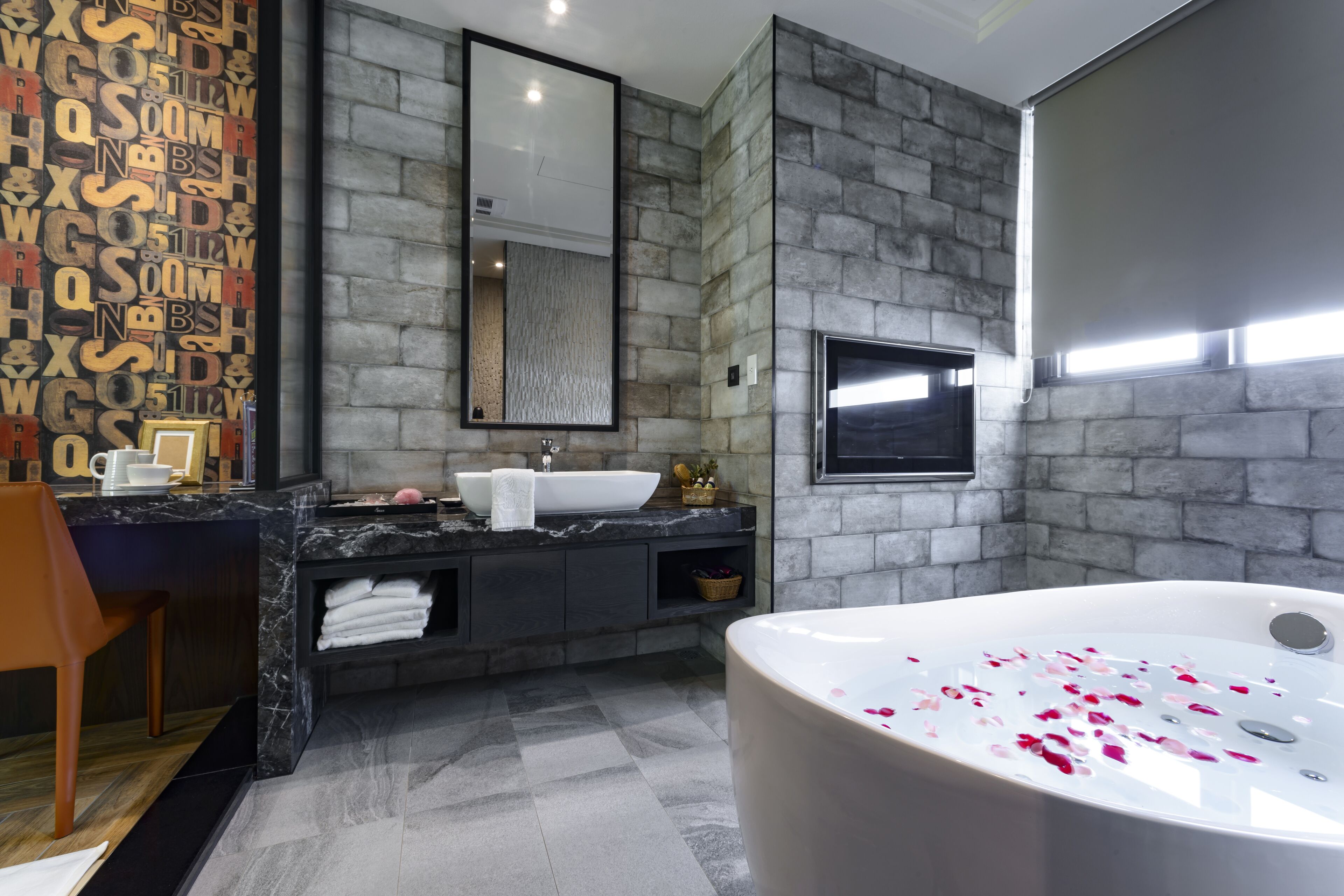 Ein modernes Badezimmer mit Steinwand, großem Spiegel, Waschbecken, einer Badewanne mit Rosenblüten und einem Holzstuhl.