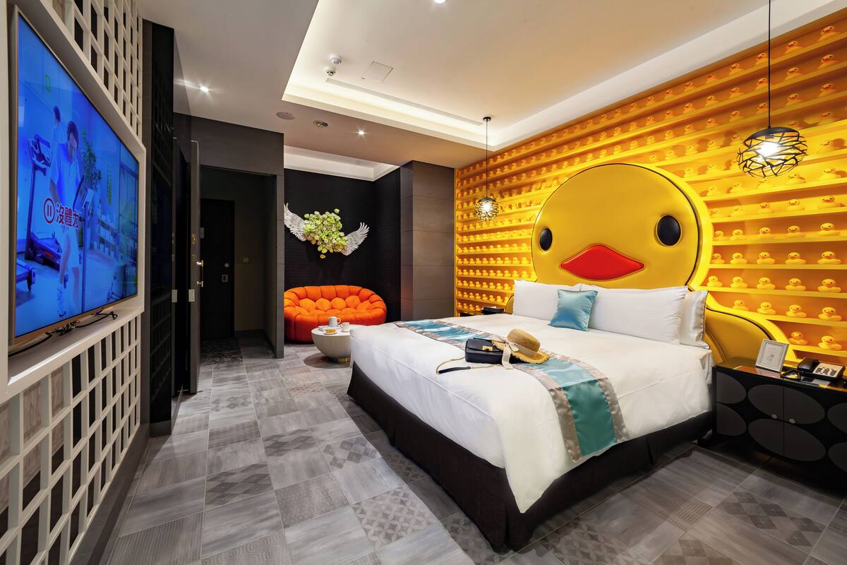 Una habitación de hotel moderna con una cama grande, televisión, un original mural con motivos de patos y un sillón naranja brillante.