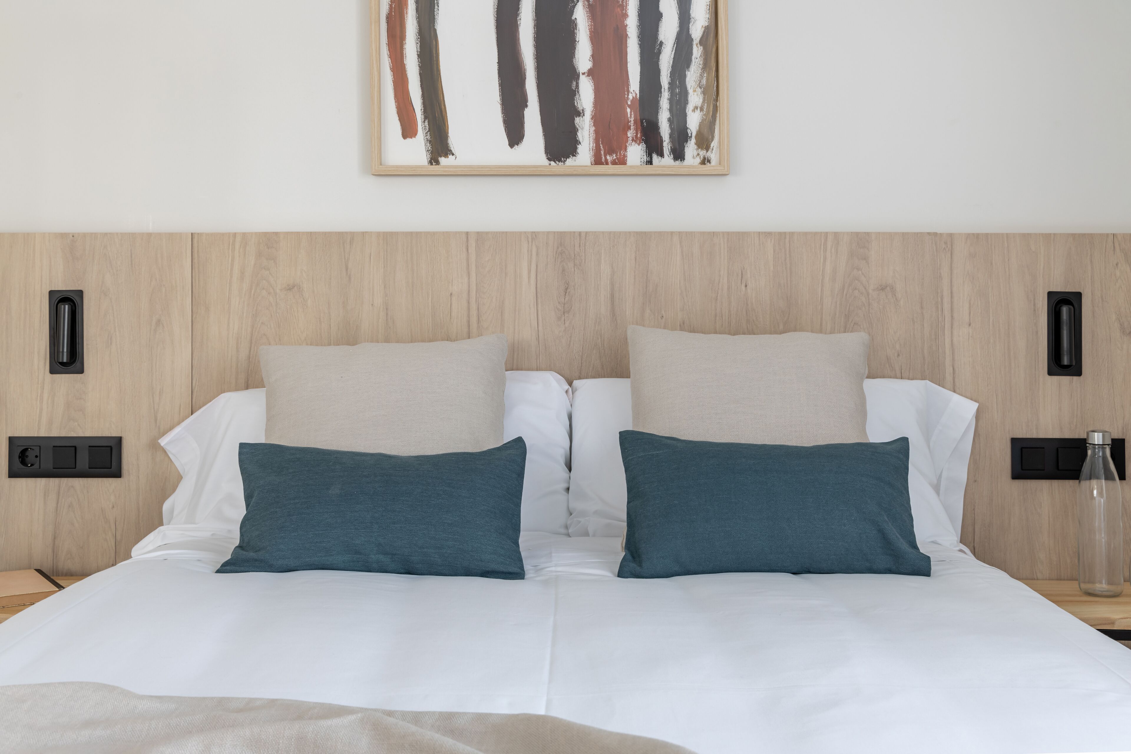 Un letto rifatto con cuscini bianchi e color acquamarina, una testiera in legno e un quadro astratto appeso alla parete.