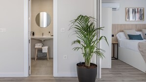 Una habitación de hotel moderna con una planta grande en maceta, una cama con ropa blanca, una mesita de noche y un aire acondicionado de pared.