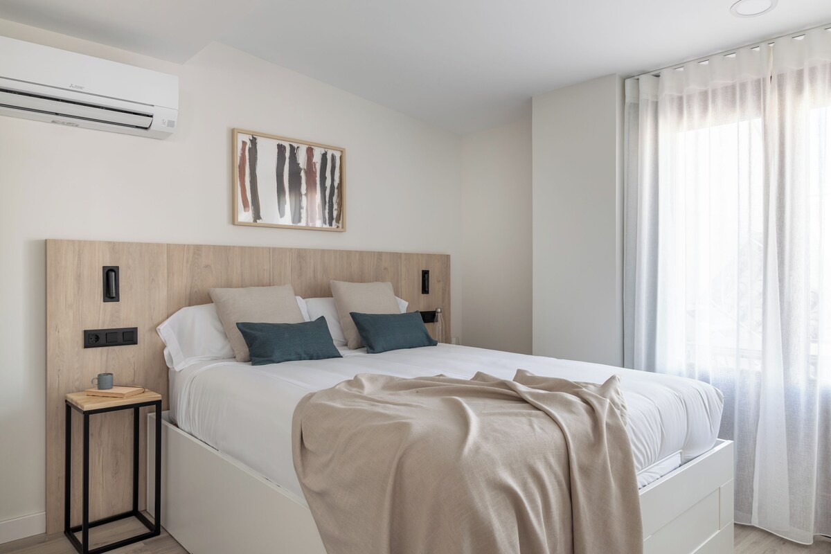 A modern bedroom with a wooden headboard, a large bed with white bedding, a bedside table with a book and mug, and a window with sheer curtains.