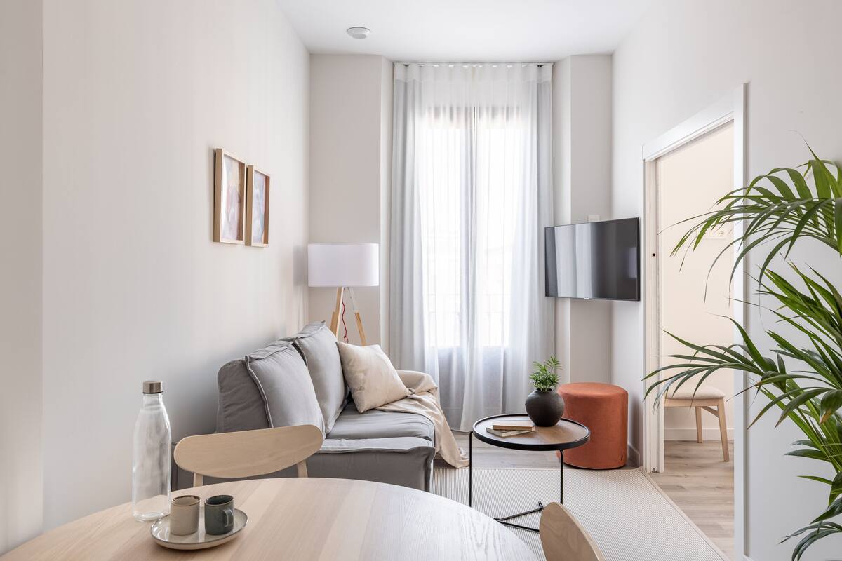 Una sala de estar moderna y minimalista con un sofá, una mesita redonda, una mesa de centro, un televisor y un ventanal con cortinas.