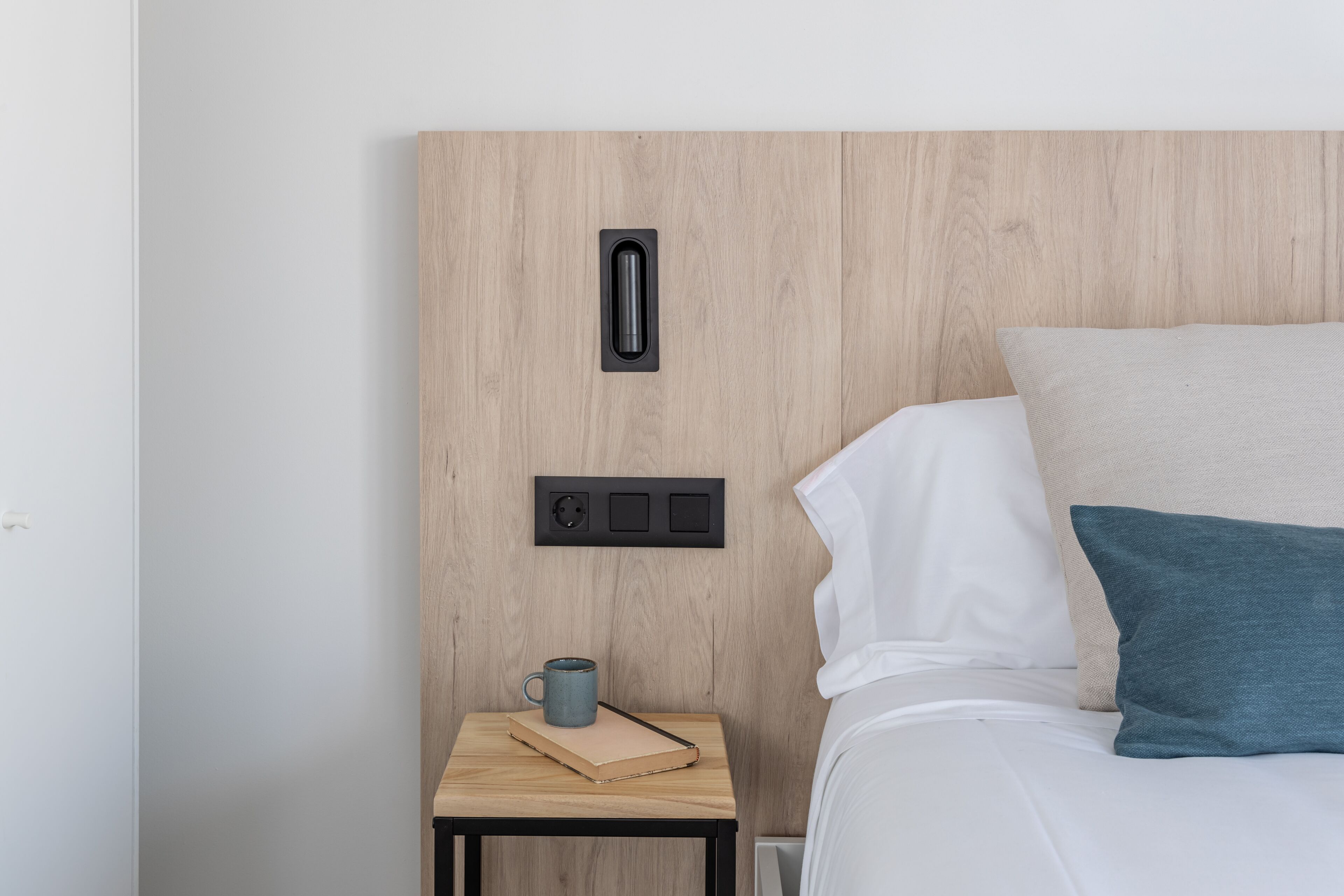 Una cama bien hecha con almohadas blancas y azul verdosas, una mesita de noche de madera con una taza y un libro, y un enchufe en la pared.