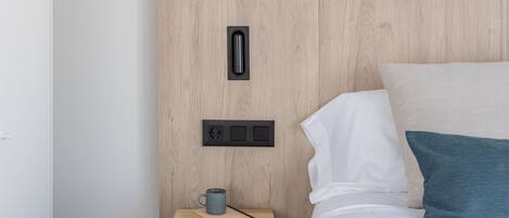 Una cama bien hecha con almohadas blancas y azul verdosas, una mesita de noche de madera con una taza y un libro, y un enchufe en la pared.