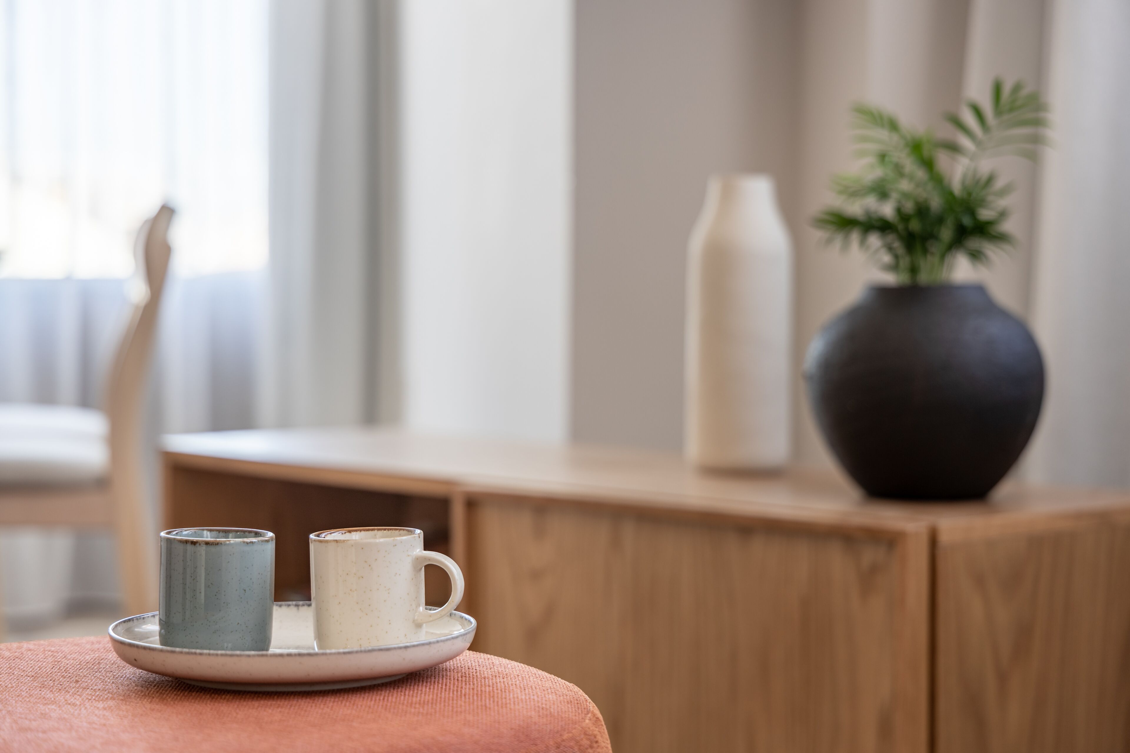 Una mesa de madera con un plato que sostiene dos tazas de cerámica, un florero con follaje verde y un jarrón blanco alto.