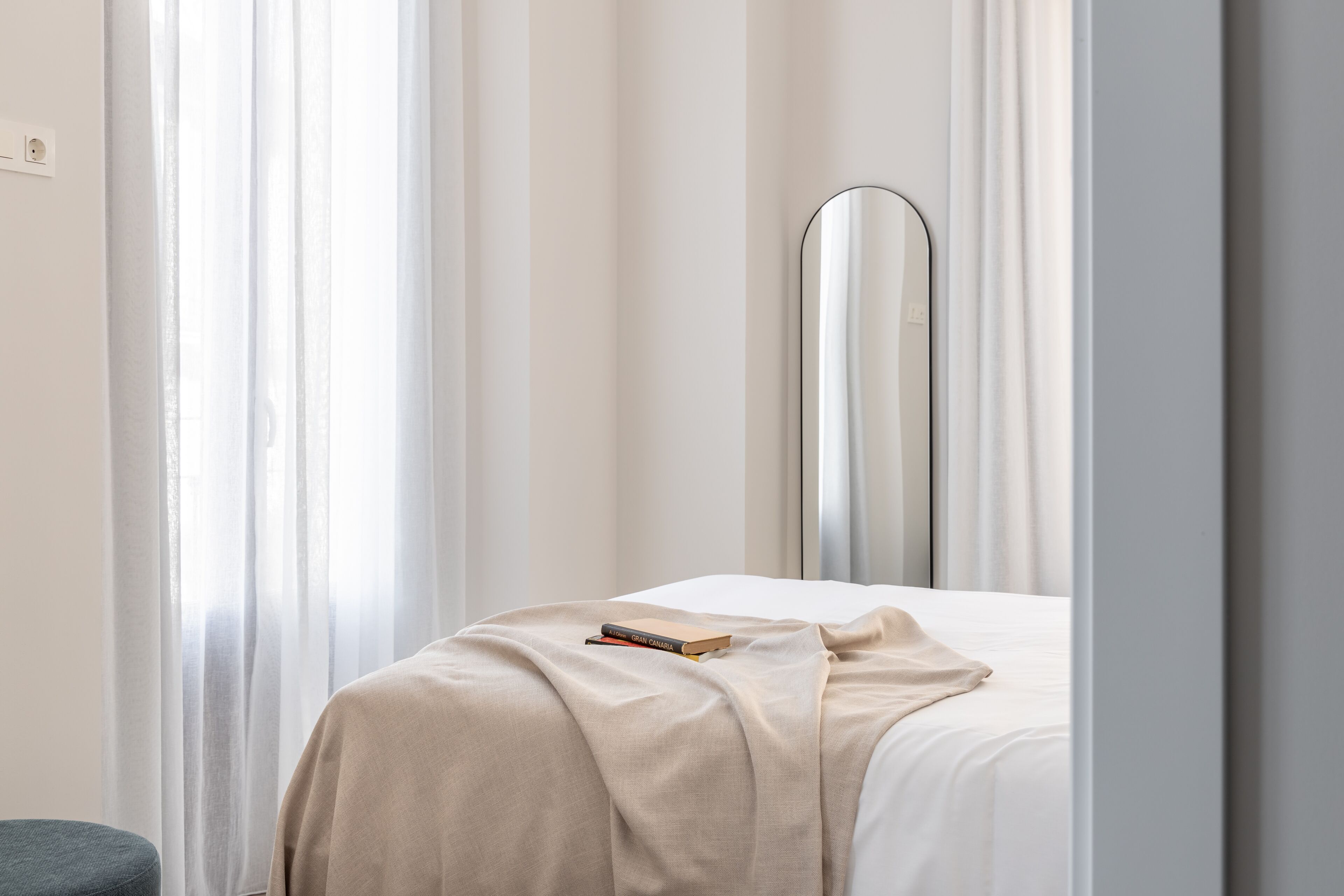 Een net opgemaakt bed met een beige deken en een boek erop, een spiegel naast het bed, en dunne gordijnen.