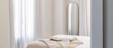 Una cama bien hecha con una manta color beige y un libro encima, un espejo en la mesita de noche y cortinas traslúcidas.