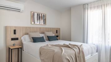 A modern bedroom with a wooden headboard, a large bed with white bedding, a bedside table with a book and mug, and a window with sheer curtains.