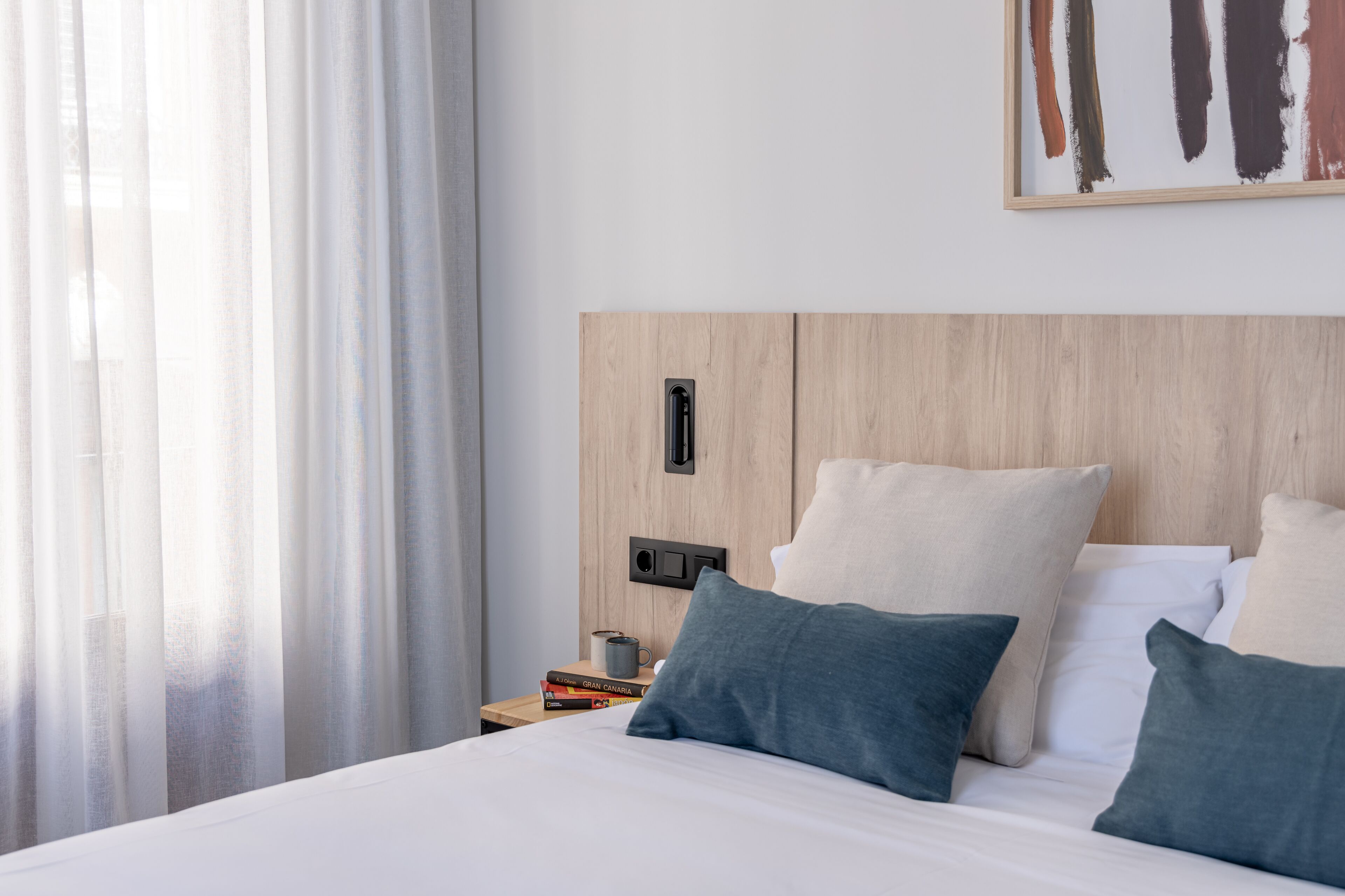 A hotel room with a wooden headboard, a bed with white and blue pillows, a nightstand with books and a mug, and a painting on the wall.