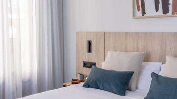 A hotel room with a wooden headboard, a bed with white and blue pillows, a nightstand with books and a mug, and a painting on the wall.