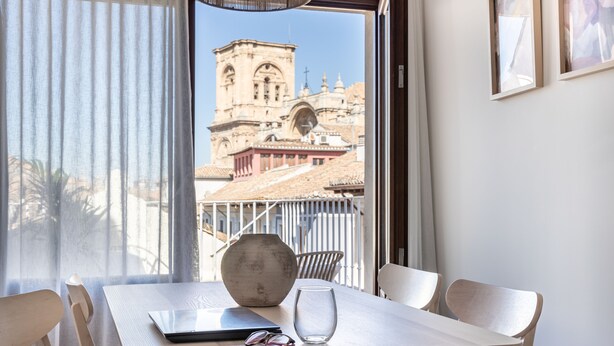 Un comedor con una mesa, sillas, un florero y una computadora portátil. Un ventanal ofrece vistas de un edificio histórico con una torre del campanario.