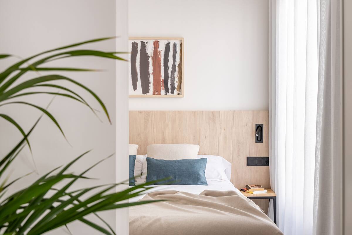 Una habitación de hotel con cabecera de madera, una cama con ropa de cama blanca y azul, una mesita de noche con un libro y un teléfono, y un cuadro en la pared.