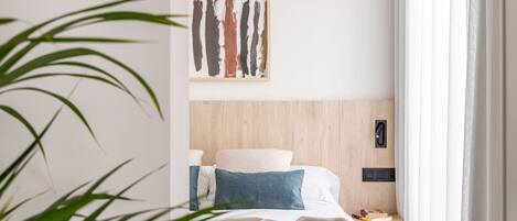 Una habitación de hotel con cabecera de madera, una cama con ropa de cama blanca y azul, una mesita de noche con un libro y un teléfono, y un cuadro en la pared.