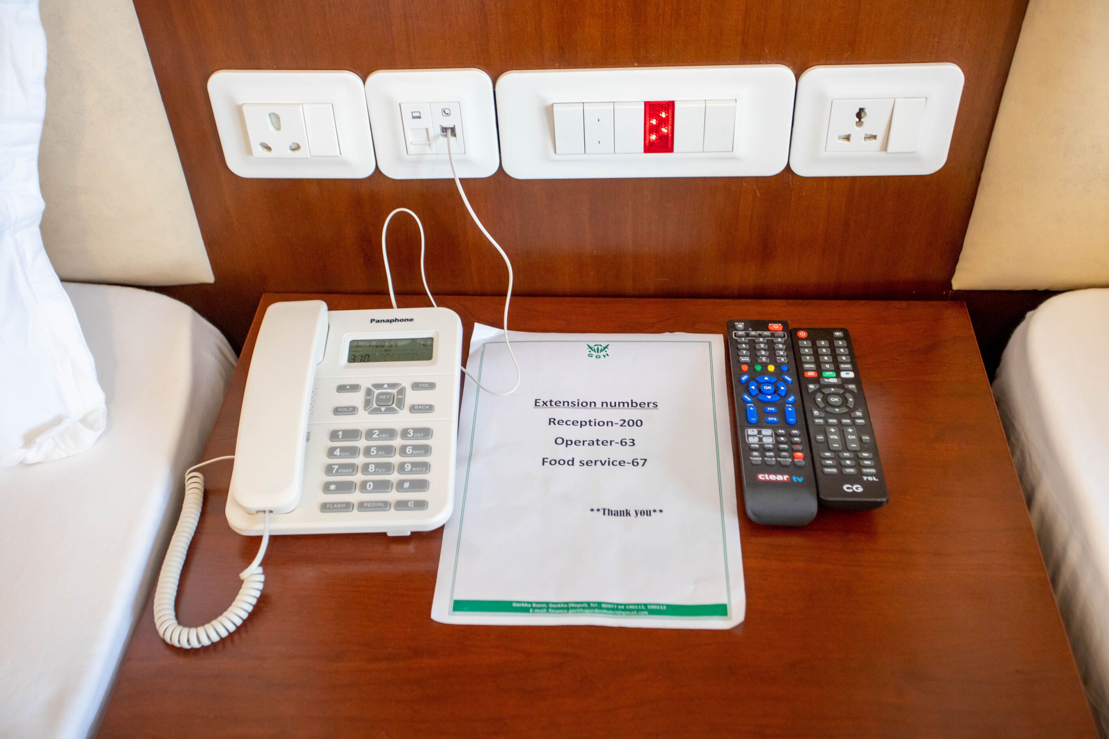 Habitación de hotel con teléfono en la mesita de noche, control remoto y un papel con números de extensión.