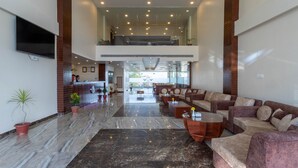 A spacious lobby with a high ceiling, a reception desk, multiple seating areas, and a large TV screen.
