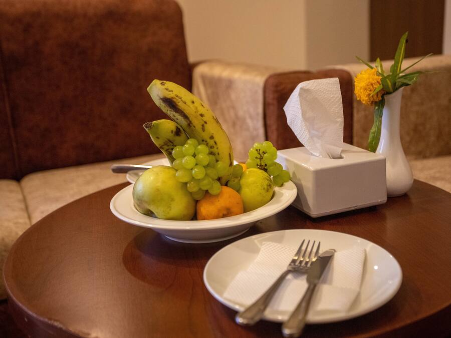 Una mesa de comedor con un frutero que contiene plátanos, uvas y manzanas, una caja de pañuelos y un plato con un tenedor y servilleta.