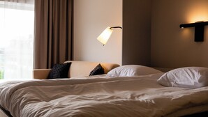 A neatly made bed with white linens, a beige headboard, and a black and white pillow. A wall-mounted lamp provides soft lighting. - Story Living Lidingö (Lidingo)