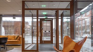 A modern lobby with a glass door entrance, a sofa, a chair, and a table with a decorative bowl. - Story Living Lidingö (Lidingo)