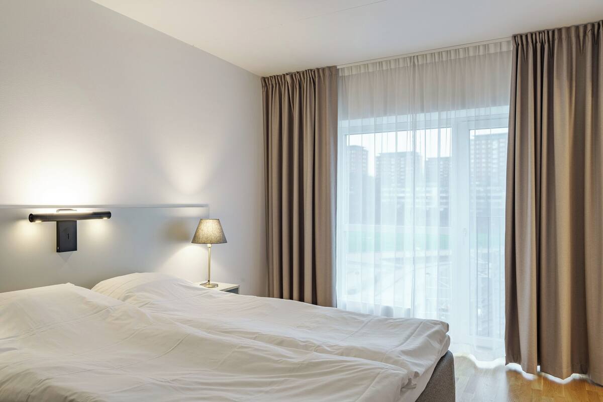 A hotel room with a large bed, a bedside table with a lamp, and a view of the city through sheer curtains.