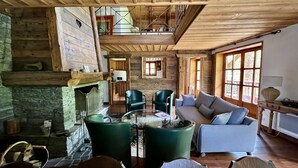 Interior rústico de una cabaña con chimenea de piedra, vigas de madera, un sillón verde, un sofá y una mesa de comedor.