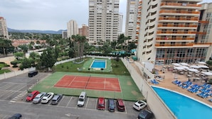 Una cancha de tenis en la azotea rodeada de edificios altos, una piscina y un estacionamiento.