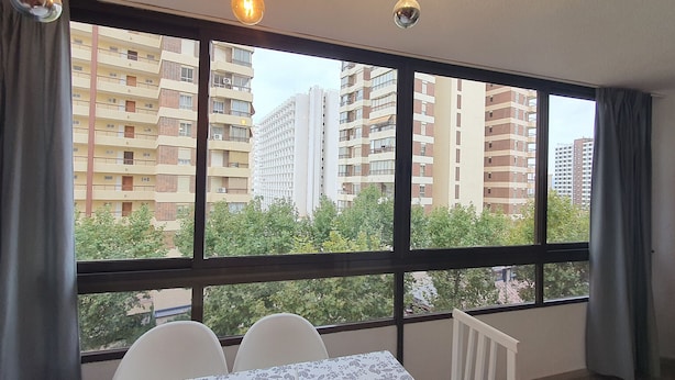 Una habitación con un ventanal grande, una mesa con sillas y vistas a edificios altos y árboles.
