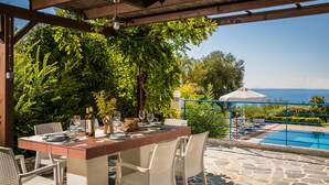 A covered outdoor dining area with a pool view, stone flooring, and white wicker chairs. - Villa Rudi (Argostoli)