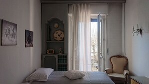 Ein Schlafzimmer mit einem Bett, einem Sessel, einem Schminktisch, einer Uhr und einem Fenster mit Vorhängen.