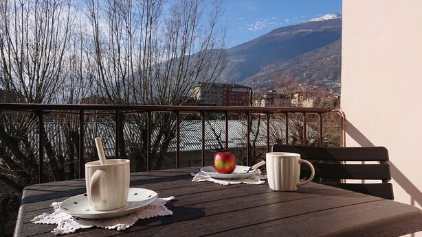 Ein Holztisch mit zwei weißen Keramiktassen und Untertellern, einem roten Apfel und einer weißen Tasse auf einem Balkon mit Blick auf eine Berglandschaft.