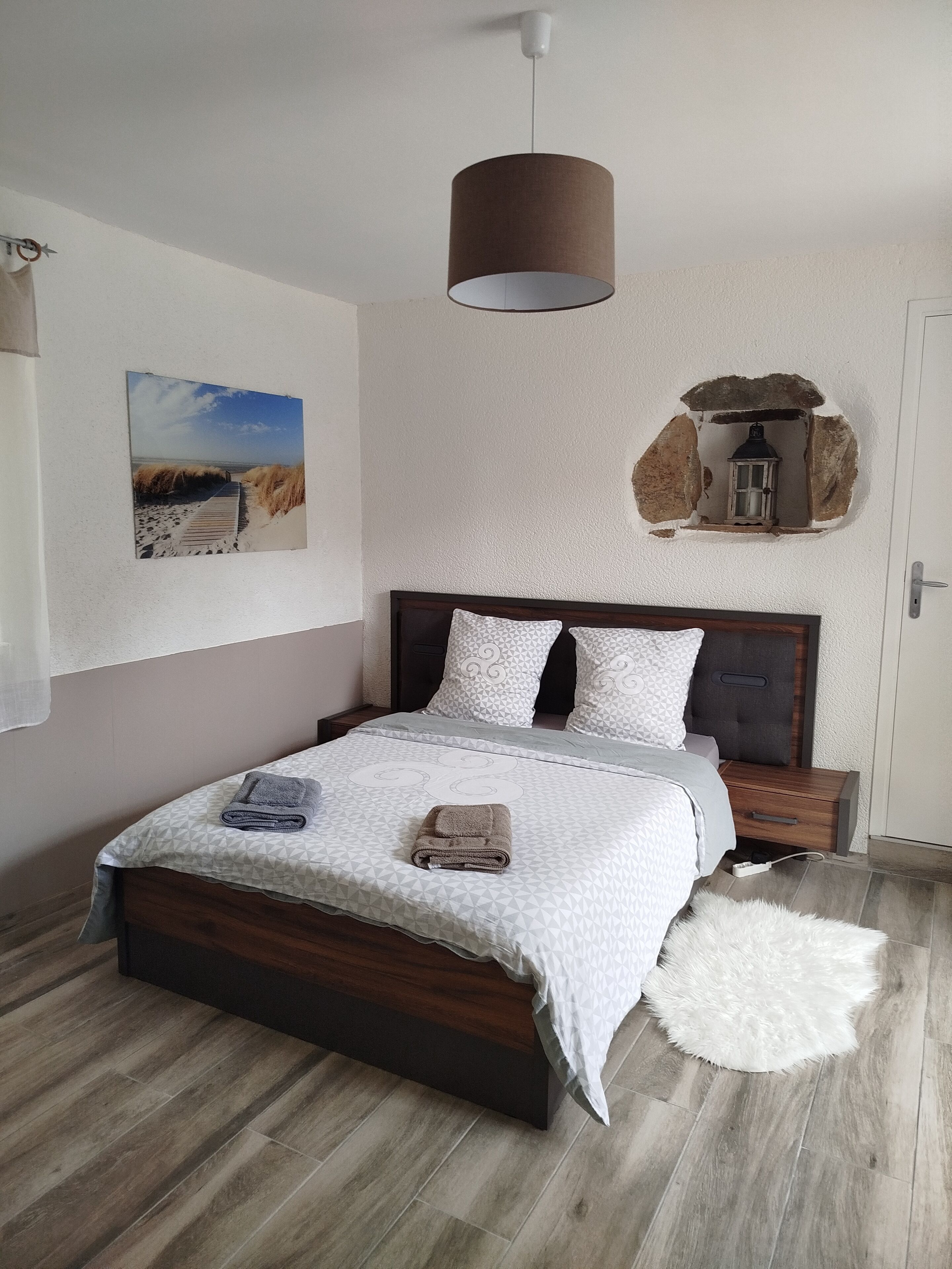 A bedroom with a wooden bed, a white wall with a beach painting, a hanging lamp, and a small window with a lantern.