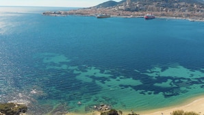 A coastal cityscape with a clear view of the sea, a prominent mountain, and a harbor with several ships.