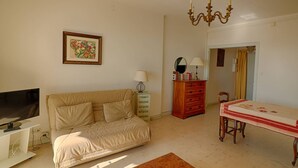 A living room with a beige sofa, a TV, a wooden coffee table, a dining table with chairs, a chandelier, and a dresser with drawers.
