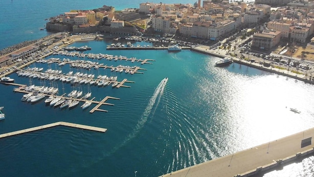 An aerial view of a coastal city with a marina filled with boats, a prominent lighthouse, and a mix of historical and modern buildings.