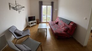 A living room with a red sofa, a TV on a white stand, a grey armchair, and a small white table. - Central Apartment with Balcony, Wi-Fi & Dishwasher - Near Shops & Spa (La Roche-Posay)