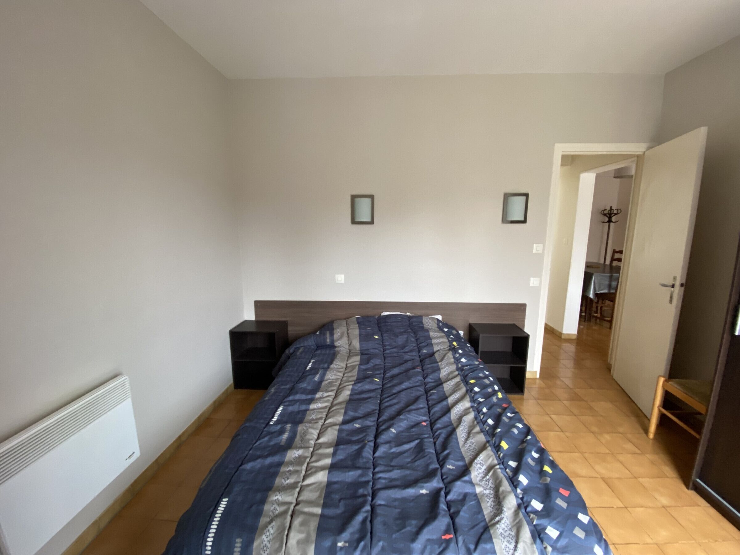 A bedroom with a bed, two bedside tables, a radiator, and a door leading to another room.