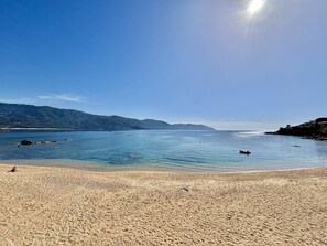 Une plage à l’eau d’un bleu limpide, bordée d’un rivage sablonneux, avec des collines qui se dessinent à l’horizon.