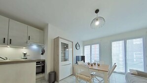 Eine moderne Küche mit weißen Schränken, ein Essbereich mit Tisch und Stühlen und ein Sofa in einem hellen Wohnraum mit großen Fenstern.