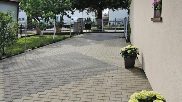 Un chemin pavé mène à une entrée fermée par un portail, l’ensemble étant entouré de verdure et bordé d’un bâtiment à la façade blanche.