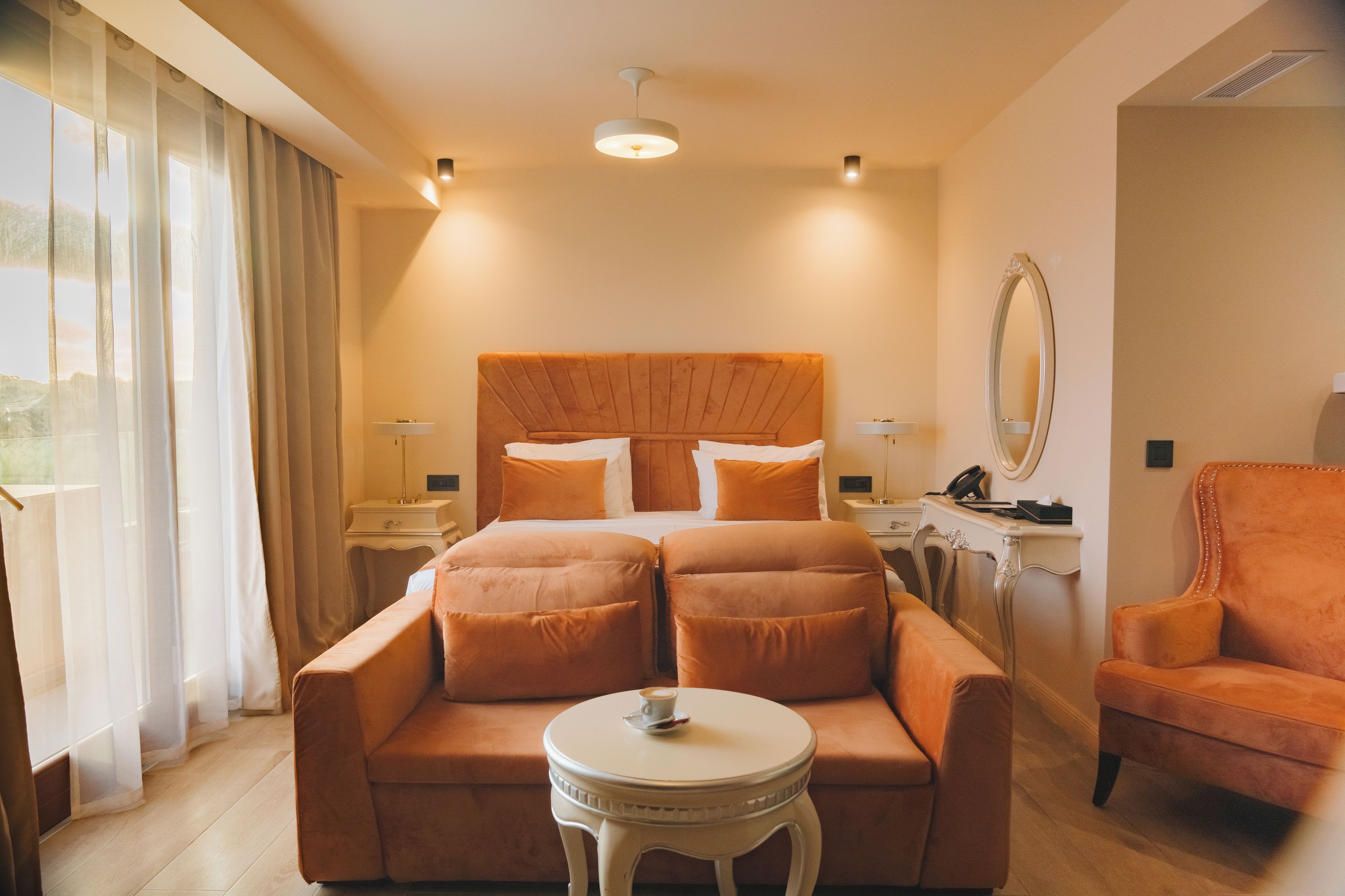 A hotel room with a large bed, a sofa, a small table with a cup, a chair, and a mirror on the wall.