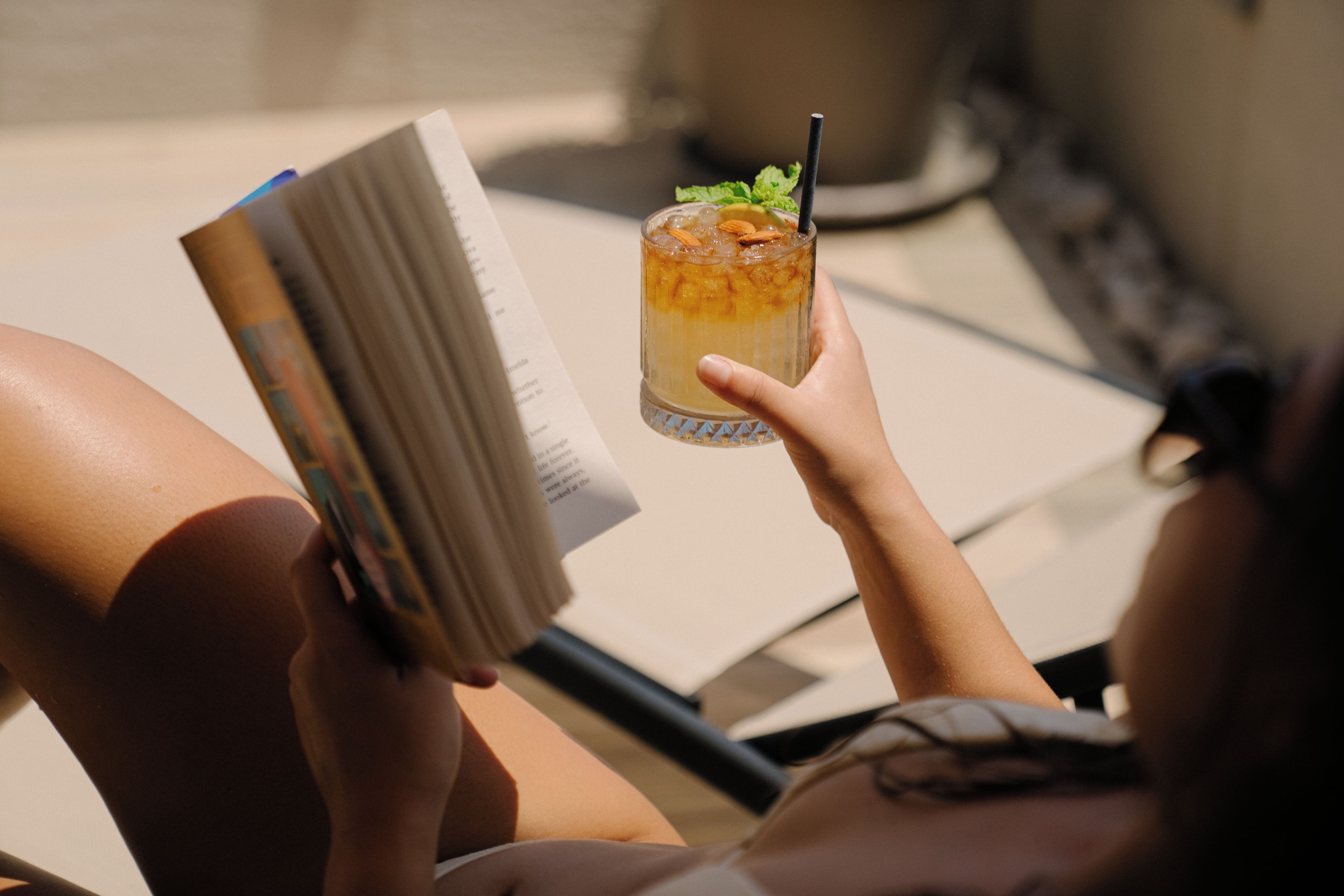A person reclining on a sun lounger, holding a book and a glass of iced beverage with a straw.