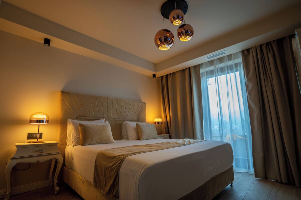 Una habitació d'hotel amb un llit gran, dues taules de nit amb làmpades, un lluminari al sostre i vistes a l'exterior a través de cortines translúcides.