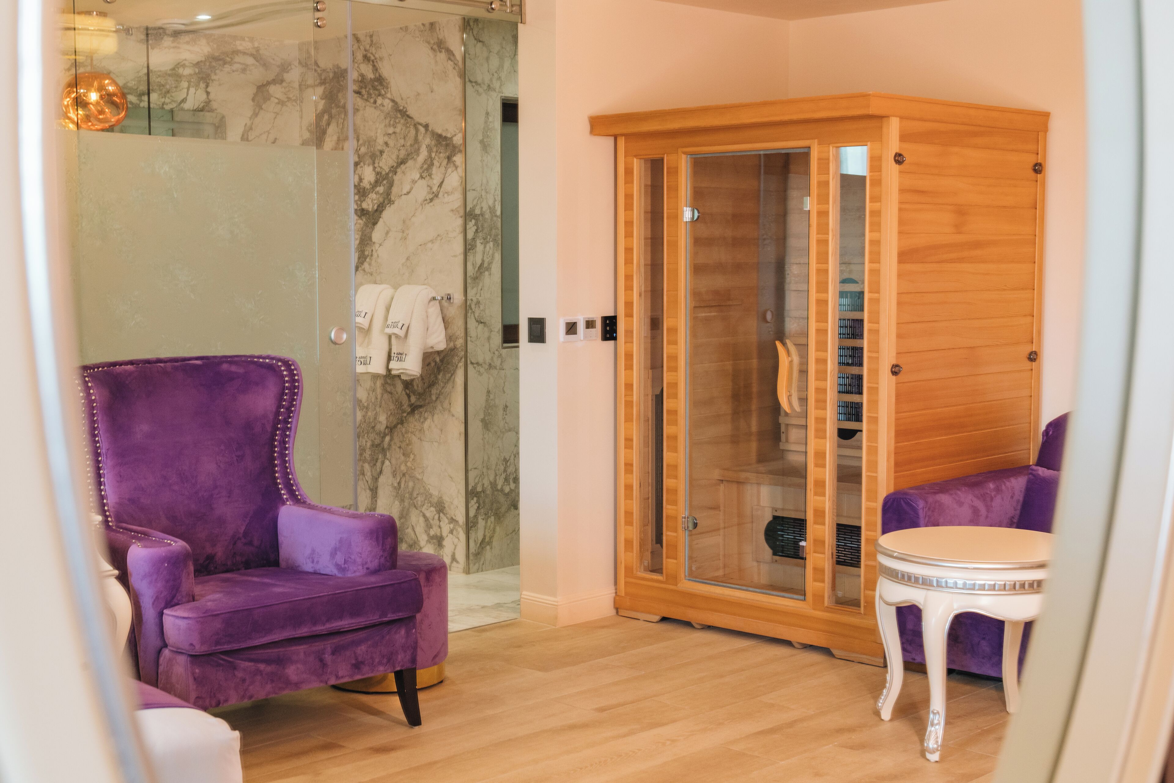 A modern bathroom with a glass shower enclosure, a purple armchair, a wooden cabinet, and a small round table.