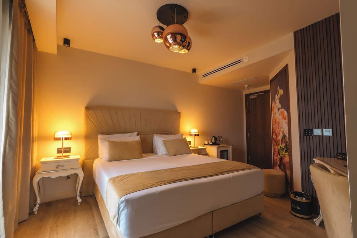 Habitació d'hotel amb un llit gran, taules de nit amb làmpades, un reposacapses, una taula de nit petita, un cistell de paper i un calefactor penjat a la paret.