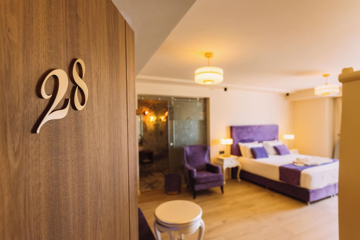 Habitació d'hotel amb una porta de fusta amb el número 28, un llit amb roba de llit morada, una taula rodona petita i un butaca morada.