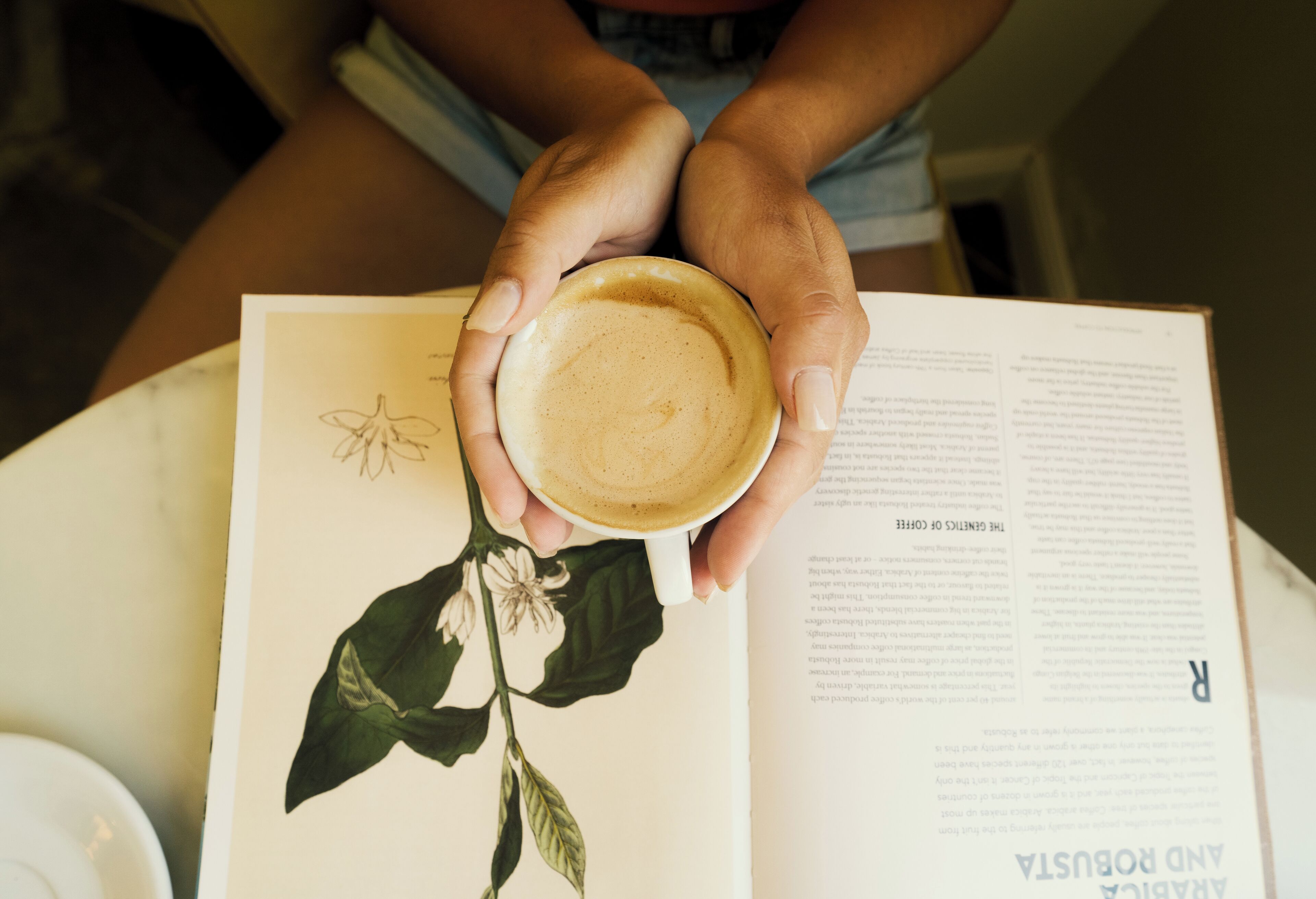 A person holding a cup of coffee with a book open beside them.