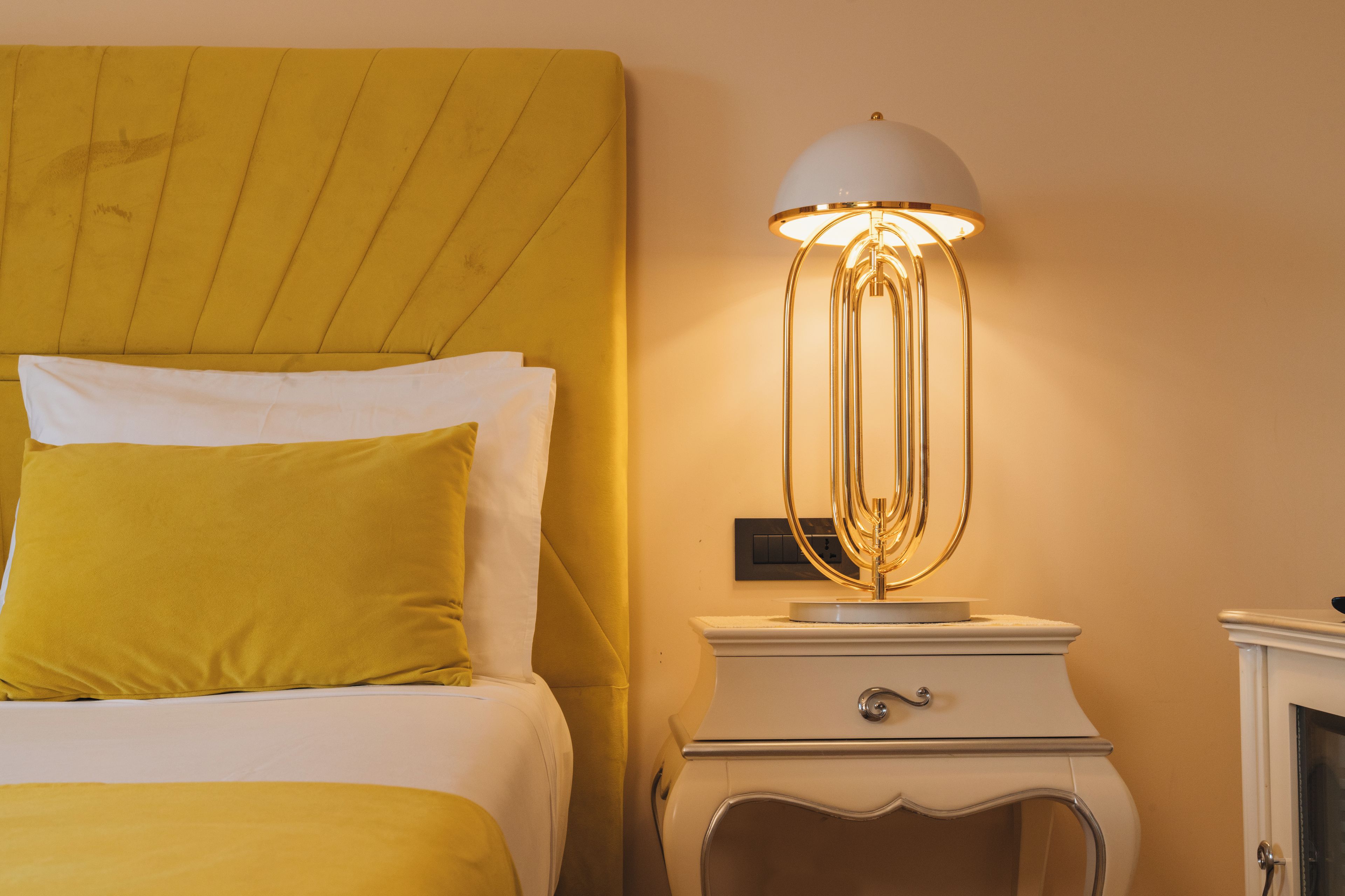 A bed with a yellow headboard and white bedding, a bedside table with a lamp, and a wall switch.